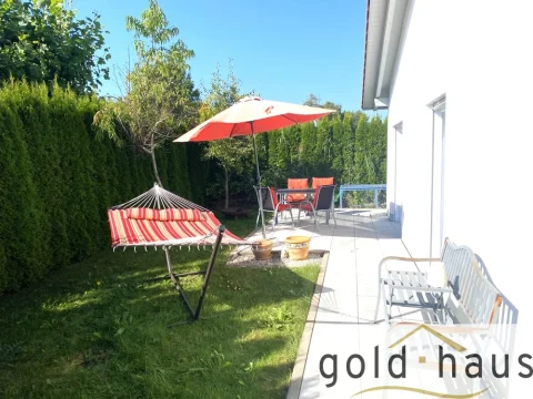 Das Bild zeigt eine sonnige Terrasse mit einem roten Sonnenschirm und einer Hängematte, Gartenmöbel mit roten Sitzkissen und einen gepflegten grünen Rasen. Umgeben ist der Bereich von hohen Hecken.