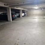 Das Bild zeigt eine beleuchtete Tiefgarage mit Betonwänden und -säulen. Zu sehen sind drei geparkte Autos: ein dunkler Kombi, ein weißes SUV und ein weiteres dunkles Auto. Auf dem Boden sind einige Flecken und zwei Kanaldeckel.