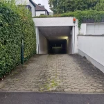 Das Bild zeigt eine Einfahrt mit einer abschüssigen Unterführung. Links ist eine große, grüne Hecke und rechts eine weiße Wand. Über der Einfahrt befindet sich ein rotes Ampellicht. Der Boden besteht aus Betonsteinen. Der String "20250816 110905" scheint ein Datum und eine Uhrzeit zu sein.