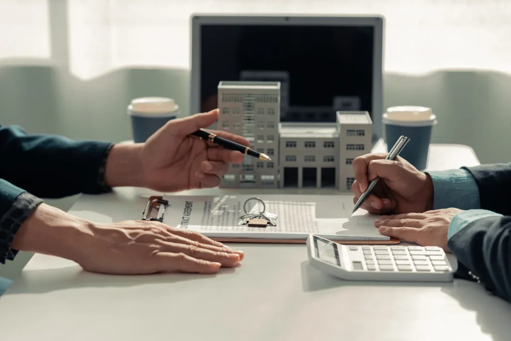 Zwei Personen sitzen an einem Tisch und diskutieren über ein Bauprojekt. Sie verwenden architektonische Modelle, Pläne, einen Taschenrechner und einen Laptop, um Details zu überprüfen.
