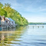 Das Bild zeigt den Ammersee mit mehreren Holzbootshäusern, die auf Pfählen im Wasser stehen. Im Hintergrund sind Berge und einige Segelboote auf dem See zu sehen. Die enge Verbindung zur Natur wird durch umgebende Bäume und klare Himmelsicht betont.