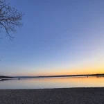Das Foto zeigt den Ammersee bei Herrsching während der Abendstimmung. Der Himmel färbt sich in sanften Blau- und Orangetönen. Kiesstrand im Vordergrund, ruhiges Wasser und ein Boot sind zu sehen. Ein Baum reicht ins Bild.