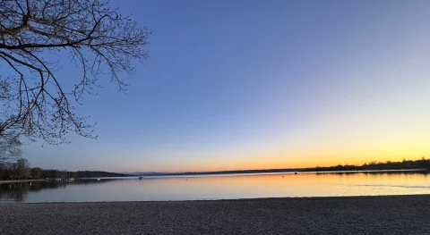 Ammersee Herrsching: Magische Abendstimmung am See Das Foto zeigt den Ammersee bei Herrsching während der Abendstimmung. Der Himmel färbt sich in sanften Blau- und Orangetönen. Kiesstrand im Vordergrund, ruhiges Wasser und ein Boot sind zu sehen. Ein Baum reicht ins Bild.