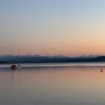 Das Bild zeigt eine ruhige Abendstimmung am Ammersee mit Blick auf die Alpen. Im Vordergrund sind mehrere Segelboote auf dem glatten Wasser zu sehen, im Hintergrund erstreckt sich die beeindruckende Silhouette der Berge, beleuchtet durch das sanfte Rosa des Sonnenuntergangs.