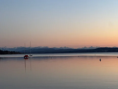 Idyllischer Sonnenuntergang am Ammersee mit Bergblick Das Bild zeigt eine ruhige Abendstimmung am Ammersee mit Blick auf die Alpen. Im Vordergrund sind mehrere Segelboote auf dem glatten Wasser zu sehen, im Hintergrund erstreckt sich die beeindruckende Silhouette der Berge, beleuchtet durch das sanfte Rosa des Sonnenuntergangs.