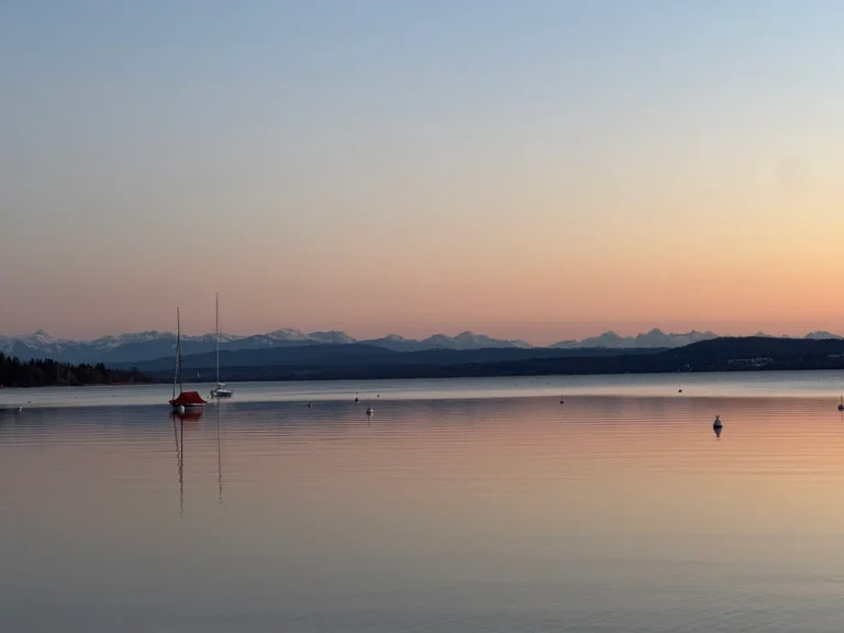 Das Bild zeigt eine ruhige Abendstimmung am Ammersee mit Blick auf die Alpen. Im Vordergrund sind mehrere Segelboote auf dem glatten Wasser zu sehen, im Hintergrund erstreckt sich die beeindruckende Silhouette der Berge, beleuchtet durch das sanfte Rosa des Sonnenuntergangs.
