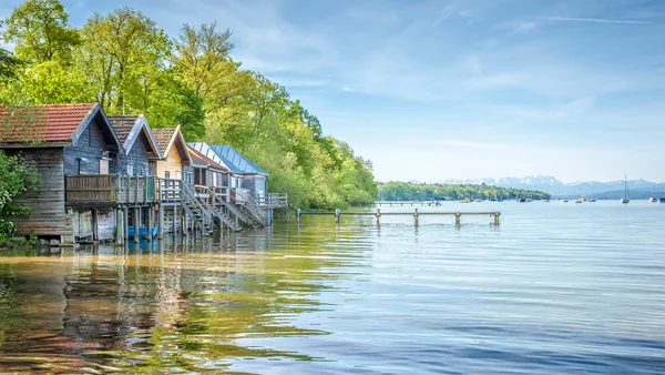 Das Bild zeigt den Ammersee mit mehreren Holzbootshäusern, die auf Pfählen im Wasser stehen. Im Hintergrund sind Berge und einige Segelboote auf dem See zu sehen. Die enge Verbindung zur Natur wird durch umgebende Bäume und klare Himmelsicht betont.