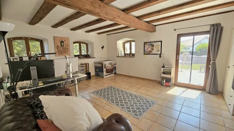 Helles Arbeitszimmer mit freiliegenden Holzbalken, zwei Schreibtischen, technischer Ausstattung und einem braunen Ledersofa. An den Wänden hängen Bilder und ein Dekor aus Korken, Terrassentür auf den Garten.