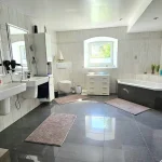 Helles, geräumiges Badezimmer mit zwei Waschbecken und großen Spiegeln, einer freistehenden Badewanne und einer separaten Duschkabine. Helles Fenster, graue Bodenfliesen und weiße Wandfliesen verleihen ein klares, modernes Aussehen.