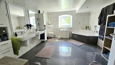 Helles, geräumiges Badezimmer mit zwei Waschbecken und großen Spiegeln, einer freistehenden Badewanne und einer separaten Duschkabine. Helles Fenster, graue Bodenfliesen und weiße Wandfliesen verleihen ein klares, modernes Aussehen.