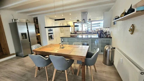 Ein moderner Essbereich in einem offenen Wohnkonzept mit angeschlossener Küche. Der Tisch aus Holz mit Platzsets und sechs Stühlen zentriert den Raum. Links eine Edelstahl-Kühlschrank. Rechts eine Küchenzeile mit Hängeschränken und Küchengeräten.