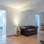 Das Bild zeigt ein helles Wohnzimmer mit einer geöffneten Tür zum weißen Badezimmer. Im Vordergrund ein dunkelbraunes Chesterfield-Sofa, daneben ein kleiner weißer Beistelltisch mit Lampe. Im Hintergrund sind ein Holzstuhl und ein Bücherregal.