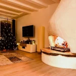 Ein gemütlicher Raum mit einem großen, gewölbten Kamin in der Ecke, in dem ein Feuer brennt. Auf der linken Seite ist ein Weihnachtsbaum mit Lichtern und Dekorationen neben einem Fernseher zu sehen. Der Boden ist mit einem großen Teppich bedeckt.