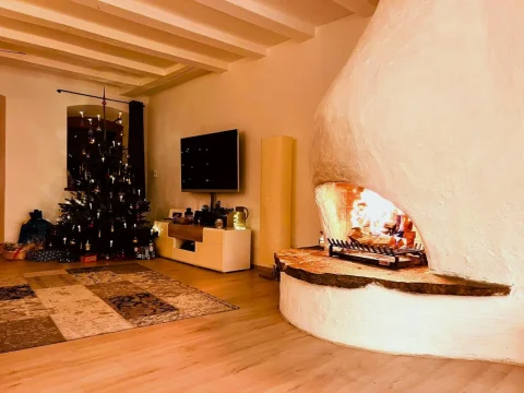 Ein gemütlicher Raum mit einem großen, gewölbten Kamin in der Ecke, in dem ein Feuer brennt. Auf der linken Seite ist ein Weihnachtsbaum mit Lichtern und Dekorationen neben einem Fernseher zu sehen. Der Boden ist mit einem großen Teppich bedeckt.