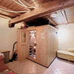 Das Bild zeigt einen Innenraum mit einer Holzsauna. Rechts befindet sich eine kleine gelbe Couch und ein runder Tisch mit einem Stuhl. Links steht eine Matratze. Überall sind Holzbalken sichtbar, was rustikales Flair vermittelt. Der Raum hat weiße Wände und Fliesenboden.