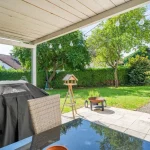 Das Foto zeigt eine Terrasse mit einem großen, abgedeckten Grill und einem Glastisch. Im Hintergrund ist ein gepflegter Garten mit Gehweg, Bäumen, einer Hecke und einem Vogelhaus auf einem Holzstativ zu sehen. Hängeleuchten zieren die Szene.