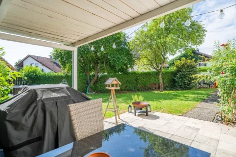 Das Foto zeigt eine Terrasse mit einem großen, abgedeckten Grill und einem Glastisch. Im Hintergrund ist ein gepflegter Garten mit Gehweg, Bäumen, einer Hecke und einem Vogelhaus auf einem Holzstativ zu sehen. Hängeleuchten zieren die Szene.