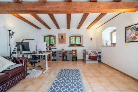 Das Bild zeigt ein helles Arbeitszimmer in einem Haus in Andechs-Frieding. Das Zimmer hat freiliegende dunkle Holzbalken und helle Wände. Ein großer Schreibtisch mit Computer, eine braune Ledercouch und viele Regale mit Büchern sind zu sehen.