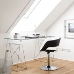 Ein moderner Arbeitsplatz unter einer Dachschräge: ein Glas-Schreibtisch mit Metallgestell, darauf ein Laptop, Smartphone, Notizen und eine Lampe. Davor steht ein drehbarer schwarzer Stuhl. Ein bodenlanges Fenster sorgt für natürliches Licht.