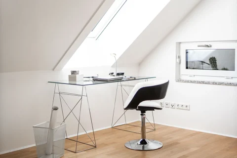 Modernes Arbeitsplatzdesign unter dem Dachgeschoss Ein moderner Arbeitsplatz unter einer Dachschräge mit einem Glas-Schreibtisch auf filigranem Metallgestell, einem schwarzen Designerstuhl, einem Laptop, einer Lampe und einer transparenten Papierkorb. Parkettboden und ein Dachfenster sind ebenfalls zu sehen.