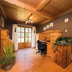 Ein gemütliches Arbeitszimmer im Tegernsee-Stil: Holzbalkendecken und -möbel, Klapptüren zum Garten, Schreibtisch mit Bürostuhl, Pflanzen und dekorative Elemente, wie Geweihe an der Wand und ein großer Fernseher.