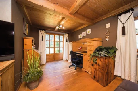 Gemütliches Arbeitszimmer Ein gemütliches Arbeitszimmer im Tegernsee-Stil: Holzbalkendecken und -möbel, Klapptüren zum Garten, Schreibtisch mit Bürostuhl, Pflanzen und dekorative Elemente, wie Geweihe an der Wand und ein großer Fernseher.