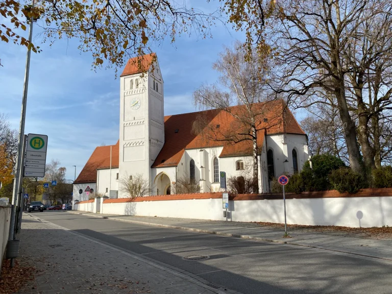 Herbstliche Szene mit einer weißen Kirche in Aubing, Sonne scheint, Laubbäume im Vordergrund.