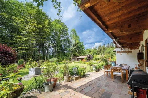 Sonnige überdachte Terrasse Landhaus Tegernsee Das Bild zeigt eine idyllische Terrasse eines Landhauses am Tegernsee mit Holzmöbeln und einem Grill. Der gut gepflegte Garten ist umgeben von hohen Bäumen. Helle, natürliche Farben dominieren die Szenerie.