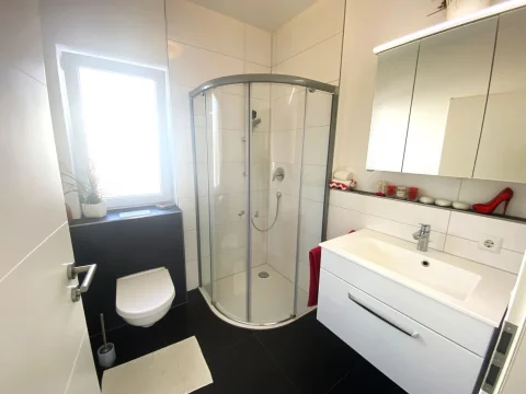 Das Bild zeigt ein modern gestaltetes Badezimmer mit einer runden Duschkabine rechts, einem weißen Waschtisch links und einer Toilette zwischen beiden. Über dem Waschtisch ist ein großer Spiegelschrank. Dekorative rote Akzente, wie ein Stöckelschuh und Handtücher, fallen auf.