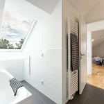 Das Bild zeigt ein Badezimmer mit einem Dachfenster durch das man Wolken sehen kann. Auf der linken Seite ist eine weiße Badewanne mit einem Handtuch. Rechts führt eine Tür zu einem Schlafzimmer mit Parkettboden.