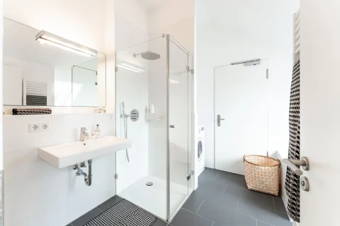 Modernes Badezimmerdesign ohne Wanne Modernes Badezimmer mit großer, gläserner Duschabtrennung, weißem Waschbecken, Spiegel mit Beleuchtung, grauen Bodenfliesen und einer Waschmaschine. Ein Korb und ein Heizkörper sind ebenfalls zu sehen. Keine Badewanne vorhanden.