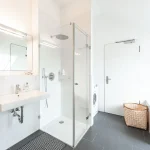 Das Bild zeigt ein modernes Badezimmer ohne Badewanne. Es enthält eine große, begehbare Dusche mit gläsernen Türen, ein längliches Waschbecken, einen Spiegel mit Beleuchtung, eine weiße Tür, eine Waschmaschine und einen Heizkörper mit einem darauf hängenden Handtuch.