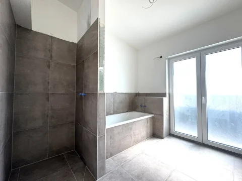 Das Bild zeigt ein modernes Badezimmer im Obergeschoss mit großen, grauen Fliesen. Eine weiße Badewanne ist in einer Ecke eingelassen, daneben eine verglaste Tür, die vermutlich auf einen Balkon oder eine Terrasse führt. Der Raum ist hell und noch nicht vollständig eingerichtet.