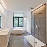 Moderne, hell beleuchtete Badsanierung mit freistehender Badewanne, bodenebener Dusche und langer Waschtischanlage. Große Fenster bieten Blick ins Grüne, dunkle Rahmen kontrastieren mit hellen Wänden und Bodenfliesen. Geschmackvoll und elegant.