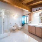 Das Bild zeigt ein Badezimmer im alpenländischen Stil, möglicherweise in einer Unterkunft am Tegernsee. Es enthält eine Duschkabine, zwei Toiletten und ein Bidet. An der Wand sind holzverkleidete Schränke und ein großer Spiegel mit Schriftzug "RELAX".