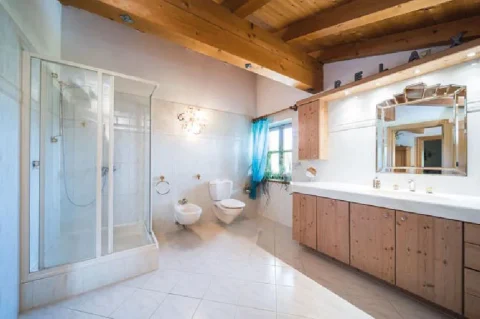 Duschbad mit Potenzial Das Bild zeigt ein Badezimmer im alpenländischen Stil, möglicherweise in einer Unterkunft am Tegernsee. Es enthält eine Duschkabine, zwei Toiletten und ein Bidet. An der Wand sind holzverkleidete Schränke und ein großer Spiegel mit Schriftzug