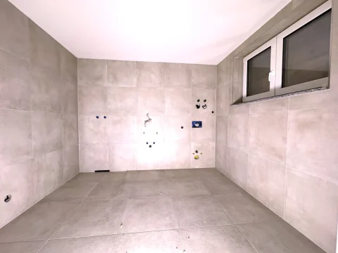 Das Bild zeigt einen unfertigen Raum, wahrscheinlich ein Badezimmer im Bau (Bad UG). Großformatige, beige Fliesen bedecken Boden und Wände. Diverse Installationsvorbereitungen wie Wasseranschlüsse und elektrische Auslässe sind sichtbar.