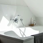 Das Bild zeigt eine helle, moderne Badewanne unter einer Dachschräge. An der Wanne ist eine verchromte Armatur mit Duschkopf befestigt. Rechts an der Wanne stehen Pflegeprodukte und ein Schwamm. Ein Korb vor der Wanne betont ein entspannendes Badevergnügen.