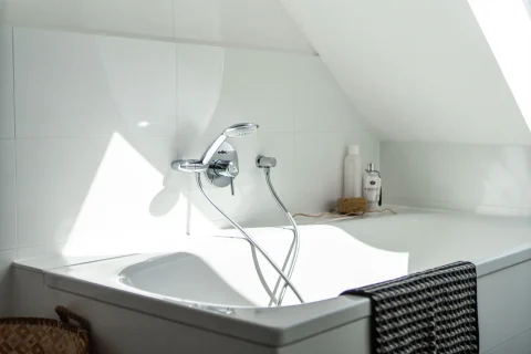 Helles Badezimmer bereit für entspanntes Badevergnügen Das Bild zeigt eine helle, moderne Badewanne unter einer Dachschräge. An der Wanne ist eine verchromte Armatur mit Duschkopf befestigt. Rechts an der Wanne stehen Pflegeprodukte und ein Schwamm. Ein Korb vor der Wanne betont ein entspannendes Badevergnügen.