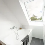 Das Bild zeigt ein helles Badezimmer mit einer weißen Badewanne. Über der Wanne ist ein großes Dachfenster, das den Blick auf den Himmel und Bäume freigibt. An der Wanne sind Armaturen mit einem Duschkopf. Neben der Wanne ist ein Korb und an der Wand hängt ein Handtuch.