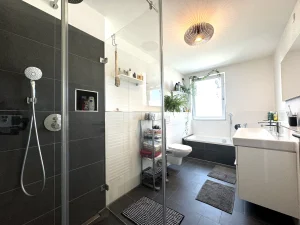 Das Bild zeigt ein helles Badezimmer mit dunklen Bodenfliesen. Eine Glasdusche, eine Badewanne unter einem Fenster, neben einer Toilette und einem Waschbecken. Dekorative Elemente sind Pflanzen und eine hängende Lampe.