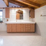 Ein rustikales Badezimmer mit hellen Fliesen, einem großzügigen Waschtisch aus Holz, einem an der Wand montierten Spiegel im Sonnenstrahlen-Design, sichtbaren Holzbalken am Dach und einem blauen Vorhang an einem kleinen Fenster.