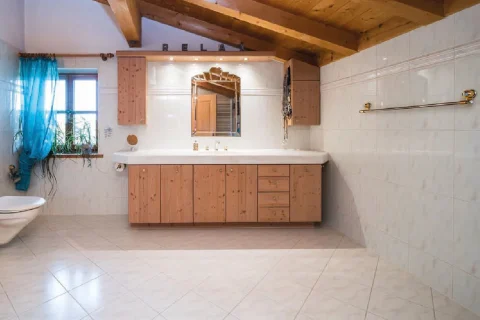 großes Badezimmer Ein rustikales Badezimmer mit hellen Fliesen, einem großzügigen Waschtisch aus Holz, einem an der Wand montierten Spiegel im Sonnenstrahlen-Design, sichtbaren Holzbalken am Dach und einem blauen Vorhang an einem kleinen Fenster.