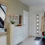 Ein moderner Eingangsbereich: Links eine weiße Kombination aus Schubladenschrank mit Spiegel darüber, auf dem Deko-Objekte liegen. Rechts, neben der Haustür, eine Garderobenstange mit Jacken und Taschen, daneben eine dunkle Treppe.
