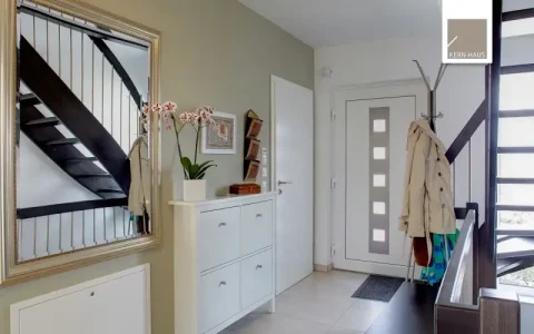 Ein moderner Eingangsbereich: Links eine weiße Kombination aus Schubladenschrank mit Spiegel darüber, auf dem Deko-Objekte liegen. Rechts, neben der Haustür, eine Garderobenstange mit Jacken und Taschen, daneben eine dunkle Treppe.