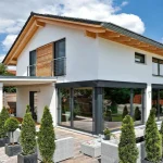 Das Bild zeigt ein modernes Einfamilienhaus in München mit weißer Fassade, Holzakzenten und Satteldach. Es besitzt eine großzügige Terrasse, umgeben von einer gepflegten Gartenanlage mit säulenförmigen Bäumen.