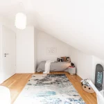 Das Bild zeigt ein minimalistisch eingerichtetes Jugendzimmer mit einer Bettnische unter einer Dachschräge. Auf dem Bett liegen mehrere Kissen und eine Wolldecke. Rechts im Bild steht ein Skateboard an der Wand. Der Raum hat einen Holzfußboden und einen gemusterten Teppich.