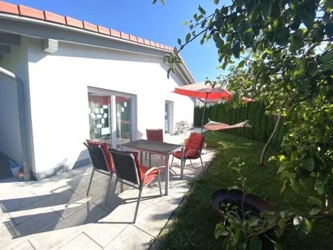 Das Bild zeigt eine sonnige Terrasse an einem Haus mit weißen Wänden und rotem Dach. Auf der Terrasse steht ein Terrassentisch mit Stühlen und einem roten Sonnenschirm, daneben eine rote Hängematte. Grüne Hecken und Bäume umgeben den Bereich.