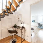 Im Vordergrund sieht man eine moderne, offene Treppe aus Metall und Holz. Links davon ein kleiner Arbeitsplatz mit Laptop und Stuhl. Durch die Türöffnung eröffnet sich der Blick in ein stilvolles Esszimmer mit großen Fenstern.