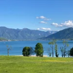 Der Blick von Gmund auf den Tegernsee zeigt eine idyllische Landschaft mit dem klaren, blauen Wasser des Sees. Im Hintergrund erheben sich grüne, bewaldete Berge. Im Vordergrund ist eine grüne Wiese mit vereinzelten Bäumen zu sehen. Ein sonniger Tag mit einigen Wolken.
